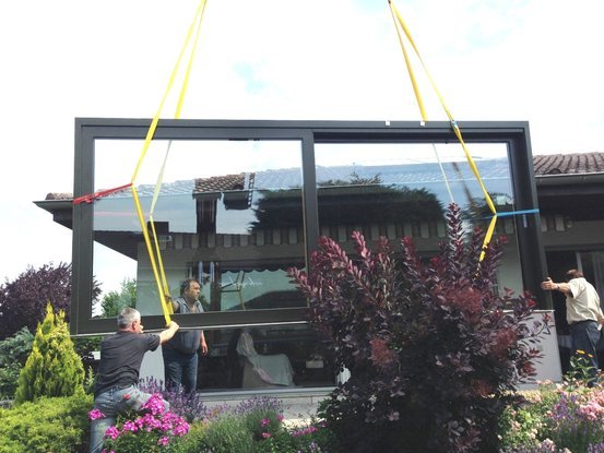 Montage einer großen Glasfront mit Hebegurten und mehreren Monteuren vor einem Haus.